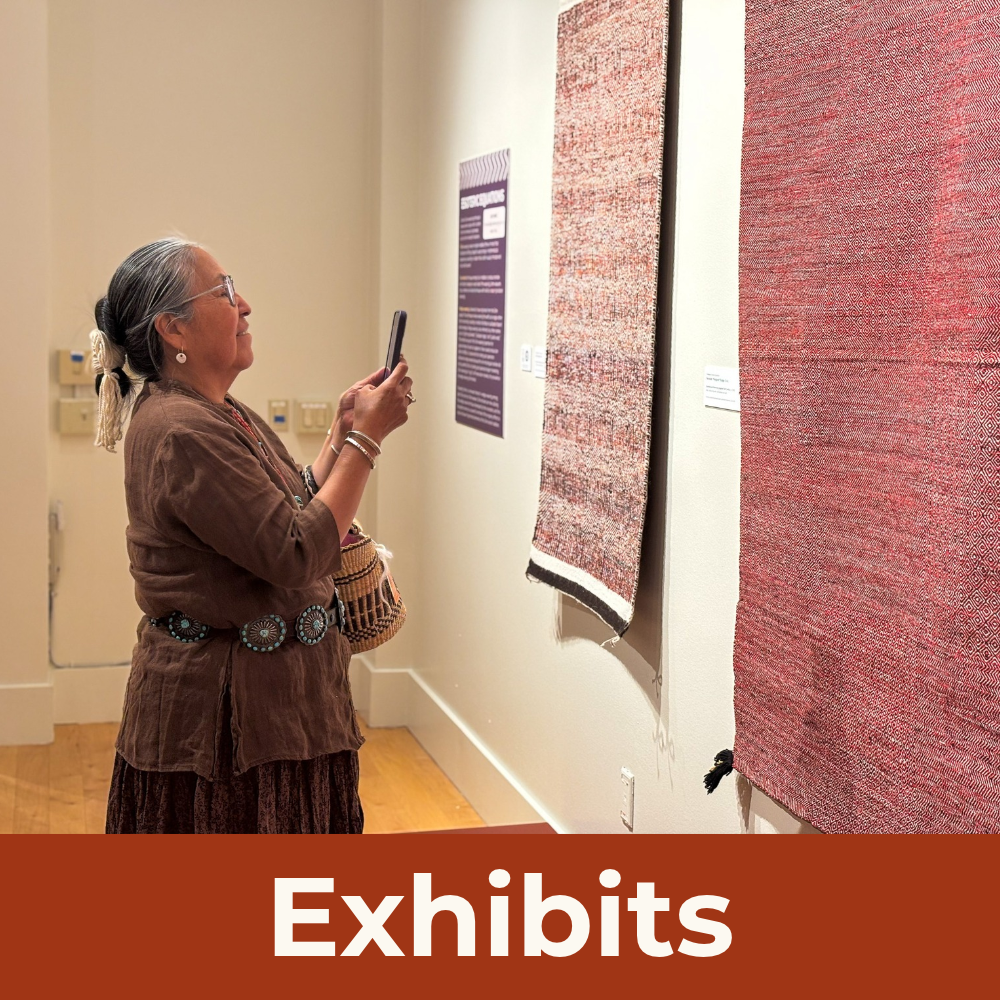 A woman stands in front of a white wall with textiles, taking a photo with her phone.